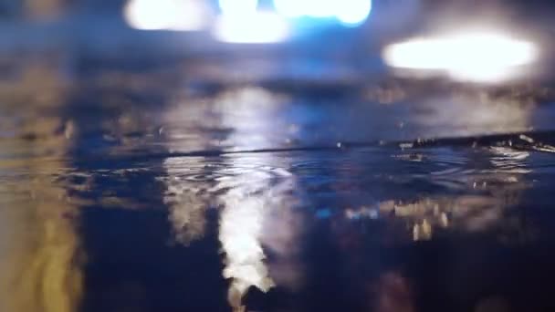 Cámara lenta primer plano gotas de lluvia cayendo en charco con luces reflexión en la acera de la ciudad . — Vídeo de stock