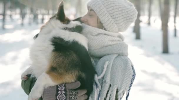 Ευτυχισμένη γυναίκα με μάλλινο καπέλο και μακρύ ζεστό μαντήλι κρατώντας, emrasing κατοικίδιο ζώο της σε χιονισμένο χειμερινό πάρκο σε παγωμένο ηλιόλουστη μέρα. Ευτυχισμένες στιγμές μαζί, χαριτωμένο σκυλί Ουαλίας Corgi Pembroke, ολομέρες χειμώνα — Αρχείο Βίντεο