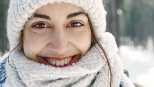 Extrém közelkép portré gyönyörű mosolygós nő gyapjú sapka és hosszú meleg sál havas téli parkban a fagyos napos napon. A nő játékosnak tűnik, és sálba burkolózik.. — Stock videók
