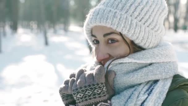 Ritratto estremo primo piano di bella donna sorridente in berretto di lana e lunga sciarpa calda nel parco invernale innevato in gelida giornata di sole. donna guarda giocoso e si avvolge in una sciarpa . — Video Stock