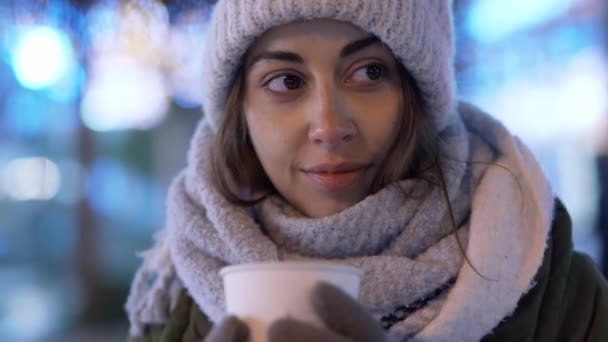 Attractive young woman in white t-shirt is sitting outdoors in park at summer evening and drinking coffee from paper cup. — Stock Video