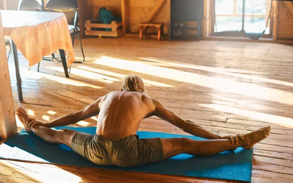 strong athletic tanned man with naked torso doing stretching at home in wooden room at sunny morning.