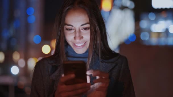 Крупный план красивой улыбающейся женщины с помощью смартфона, гуляющей по ночному городу — стоковое видео