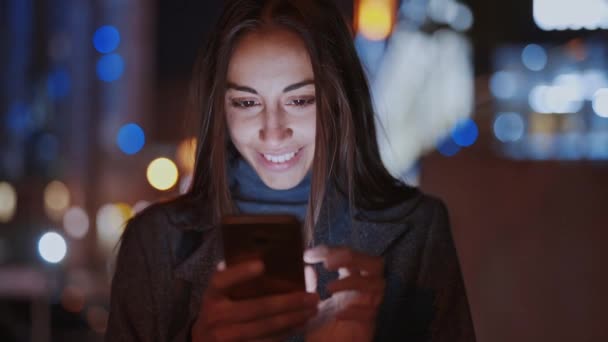 Primer plano de prety sonriendo mujer usando smartphone caminando por la noche ciudad — Vídeo de stock