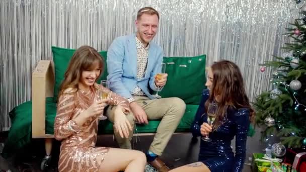 Две молодые стильные девушки и один мужчина, сидящий с бокалами шампанского на вечеринке, разговаривающий и смеющийся — стоковое видео
