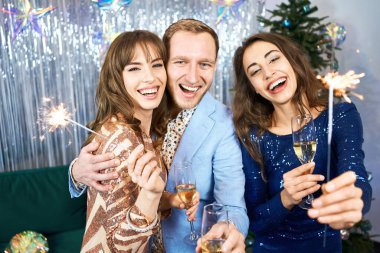 Şampanya kadehleriyle, gülen, gülümseyen ve gümüş arkaplanda maytap tutan bir grup güzel genç..