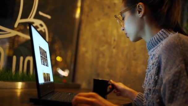 Young woman freelancer sitting at night in cafe and using laptop, looking at monitor and drinking coffee — Stock Video