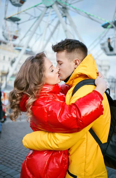 Giovane coppia romantica innamorata baciare, abbracciare, camminare per strada, indossando piumini luminosi. ruota panoramica sullo sfondo . — Foto Stock