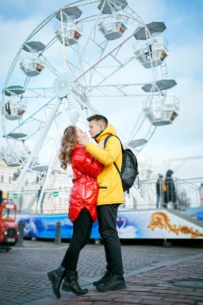 Giovane coppia romantica innamorata baciare, abbracciare, camminare per strada, indossando piumini luminosi. ruota panoramica sullo sfondo . — Foto Stock