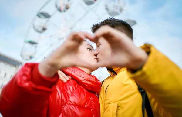 Genç ve romantik bir çift, parmaklarıyla yaptıkları bir kalp jesti ile sarılıp öpüşüyorlar, şehirde birlikte vakit geçiriyorlar. Aşıklar parlak sarı ve kırmızı ceketler giyer. — Stok fotoğraf