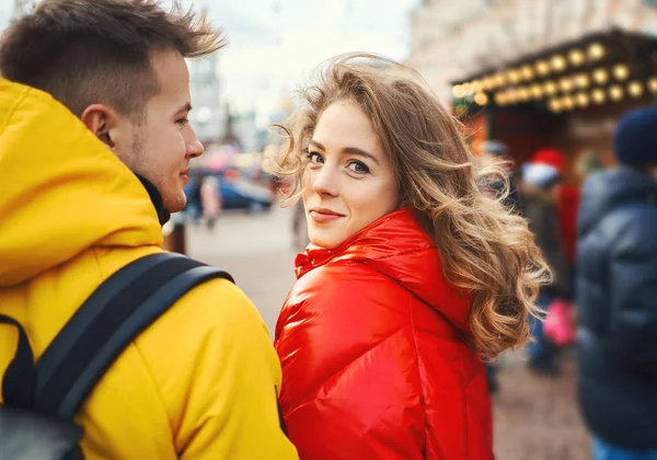 魅力的な若いです女性で赤いダウンジャケットは好転し、カメラを見て,街で彼女のボーイフレンドと一緒に歩いて. — ストック写真