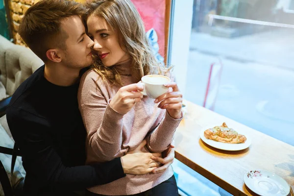 Pasangan romantis yang bahagia duduk di atas jendela di kafe, memeluk, mencium dan minum kopi. Pacar lembut memeluk pacar di kafe nyaman. — Stok Foto