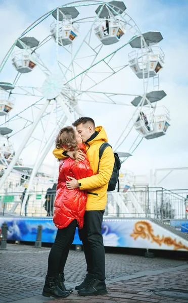 Giovane coppia romantica baciare, abbracciare, camminare per strada, indossarsi in piumini luminosi. ruota panoramica sullo sfondo. — Foto Stock