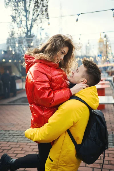 Tineri cuplu romantic în îmbrățișare dragoste, purtând în galben strălucitor și jachete roșu jos. Bărbat frumos care transportă o tânără femeie atractivă în timp ce se plimbă pe stradă . — Fotografie, imagine de stoc