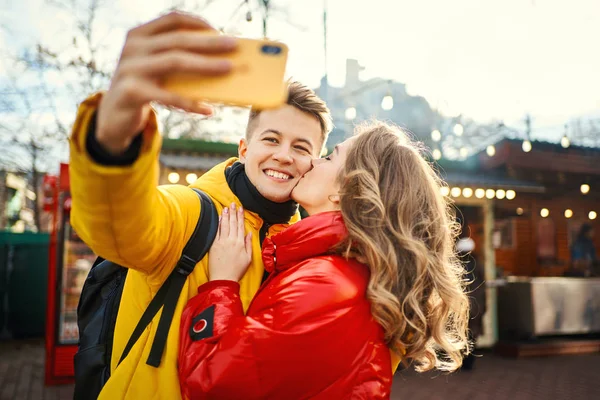 Молодая романтическая пара влюбленных делает селфи на смартфоне, улыбается камере, женщина целует в щеку своего бойфренда. — стоковое фото