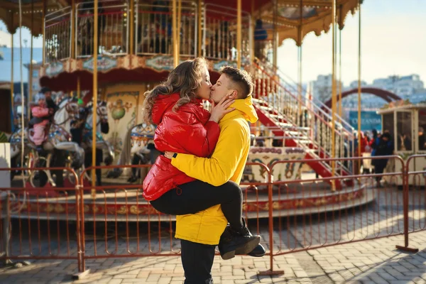 Parlak sarı ve kırmızı ceketli genç romantik çift öpüşüyor, sarılıyor. Fransız atlıkarıncasında genç ve çekici bir kadını tutan yakışıklı bir adam.. — Stok fotoğraf