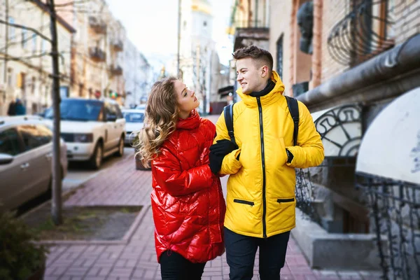 Felice giovane coppia che cammina per strada della città, guardandosi con sorriso, indossarsi in giubbotti gialli e rossi brillanti. — Foto Stock