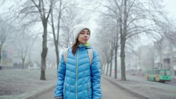 Молодая спортсменка в синей куртке и шерстяной шляпе гуляет по переулку в туманный зимний день — стоковое видео
