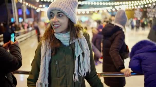 Happy smiling young woman wearing warm winter clothes standing on city ice skating rink background — Stock Video