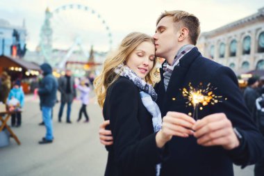 Sokakta ışıldayan ve arka planda cazibesi olan zarif romantik bir çift. Adam sevgilini kucaklıyor ve şefkatle öpüyor.