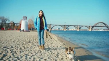Genç bir kadın deniz kıyısındaki kumlu sahil boyunca sevimli bir köpekle yürüyor.