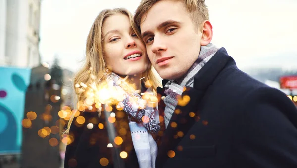 Ritratto di elegante coppia romantica sorridente alla macchina fotografica, con scintilla nelle mani su sfondo di addobbi festivi della città. — Foto Stock