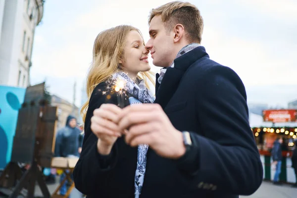 Elegante coppia bionda con scintilla tra le mani abbracciando e baciandosi su sfondo di addobbi festivi della città. — Foto Stock