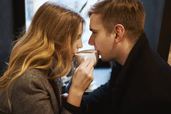 Young happy couple sitting over the window in cafe and drinking coffee from one cup. — Stock Photo, Image