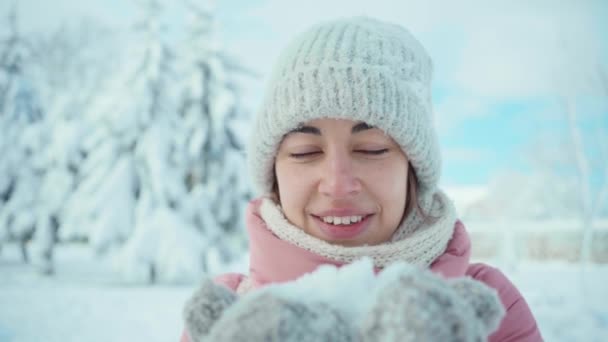 Câmera lenta bela jovem em parka rosa e chapéu de malha e luvas sopra neve das palmas das mãos — Vídeo de Stock