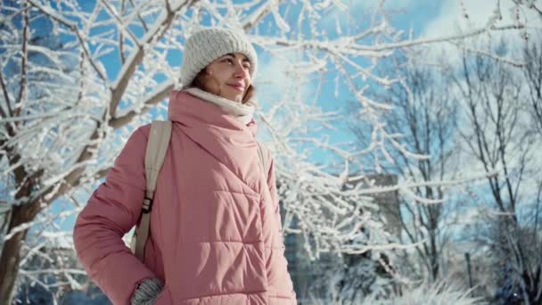 Αργή κίνηση πορτρέτο όμορφη χαμογελαστή νεαρή γυναίκα σε ζεστό ροζ μπουφάν με τα πόδια στο χιονισμένο χειμερινό πάρκο σε αφρώδη ηλιόλουστη μέρα — Αρχείο Βίντεο