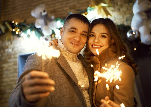 รูปภาพที่สดใสของคู่รักโรแมนติก ยิ้มกับกล้อง ด้วยแสงสว่างในมือ ยืนต่อต้านการตกแต่งวันหยุดที่สดใส . — ภาพถ่ายสต็อก