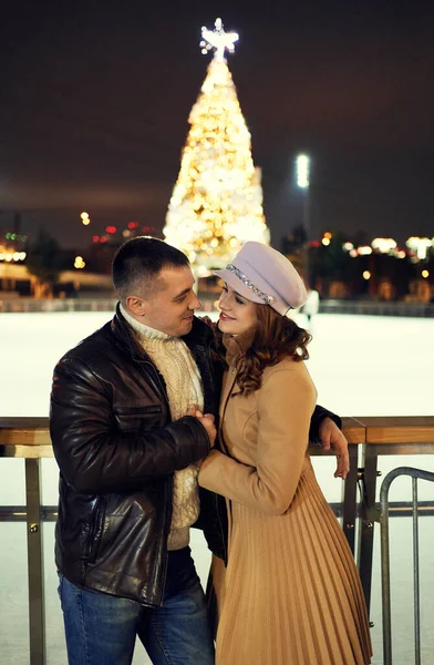 Romantic stylish couple in warm clothes embracing, looking at each other with smile, standing against ice rink with bright Cristmass tree. — Stock Photo, Image