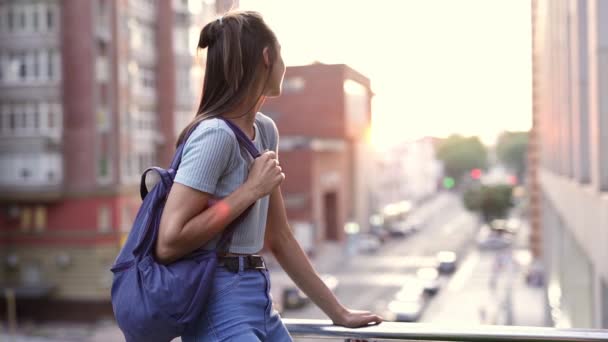 Feliz sorrindo jovem mulher em desgaste casual com pequena mochila sentada no corrimão da varanda — Vídeo de Stock