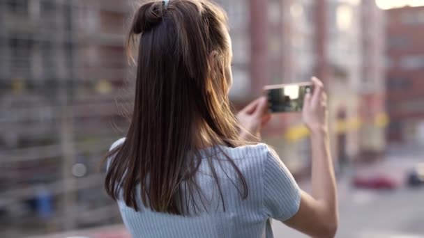 Молодая женщина, стоящая на фоне улицы Сансет Сити и снимающая фото или видео с помощью смартфона — стоковое видео
