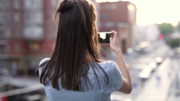 Mujer joven de pie en el fondo de la calle de la ciudad puesta del sol y tomar fotos o videos con un teléfono inteligente — Vídeo de stock