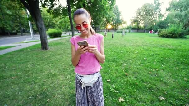 Smiling woman in pink eyeglasses standing on sunset city green park background and using mobile phone — Stock Video