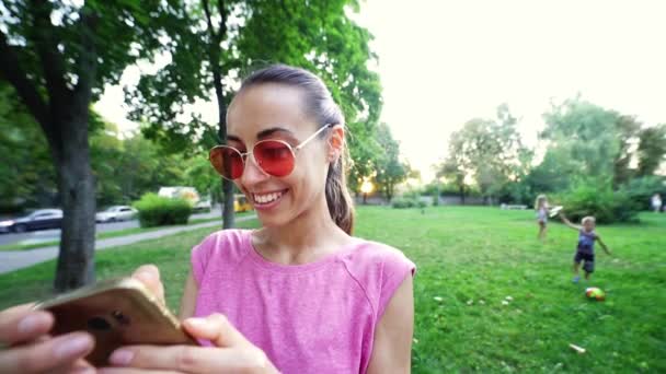 Leende kvinna med roligt ansikte och rosa glasögon står på solnedgången stad grön park bakgrund och med mobiltelefon — Stockvideo