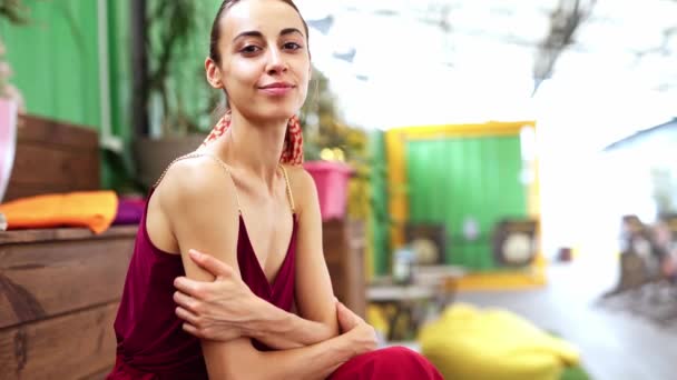 Beautiful elegant young woman in a burgundy light sundress sits on a wooden bench in a summer cafe — Stock Video