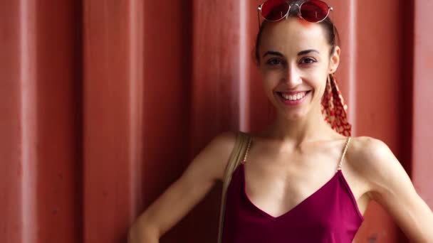 Радостная молодая женщина в бордовом сарафане позирует на фоне красной стены, смотрит на камеру и надевает розовые очки — стоковое видео