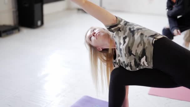 Attraente donna bionda in primo piano e gruppo di giovani sportivi che praticano yoga al coperto — Video Stock