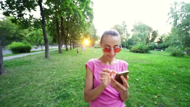 Pomalý pohyb mladá usměvavá žena v růžové brýle pomocí mobilního telefonu na západ slunce město zelený park pozadí — Stock video