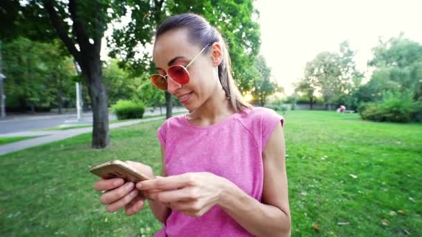 Pomalý pohyb mladá usměvavá žena v růžové brýle pomocí mobilního telefonu na západ slunce město zelený park pozadí — Stock video