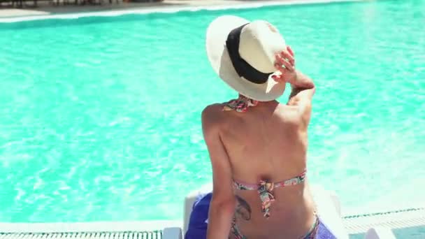 Back view of young woman in bikini and hat relaxing in desk chair on poolside background. — Stock Video