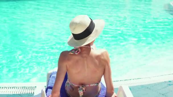 Back view of young lady in bikini and hat relaxing in desk chair on poolside background. — Stock Video