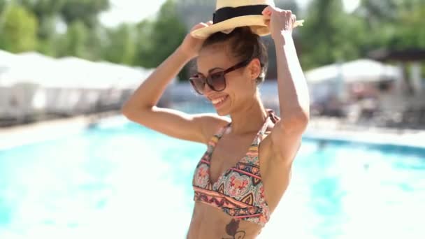 Pretty young woman in bikini, hat and sunglasses smiling joyfully at the pool background. — Stock Video