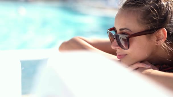 Cerca del retrato de la bella niña caucásica sonriente en gafas de sol que se encuentra en el borde de la piscina en la silla de escritorio y tomando el sol. — Vídeos de Stock