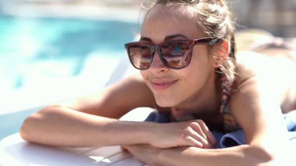 Közeli portré gyönyörű mosolygós kaukázusi lány napszemüvegben feküdt a medence szélén az íróasztal szék és napozás — Stock videók