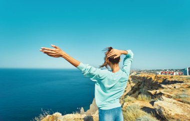 Kaygısız genç bir kadının arkası. Uçurumun kenarında duruyor. Güzel deniz manzarası ve harika doğanın tadını çıkarıyor..