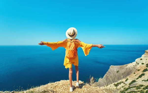 Rear view of female traveler in bright yellow dress and hat with backpack standing the top of mountain with raised hands against amazing seascape. — kuvapankkivalokuva