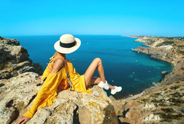 Side view of traveler woman in bright yellow dress and hat sitting on cliff edge with beautiful sea view and enjoying wonderful nature – stockfoto